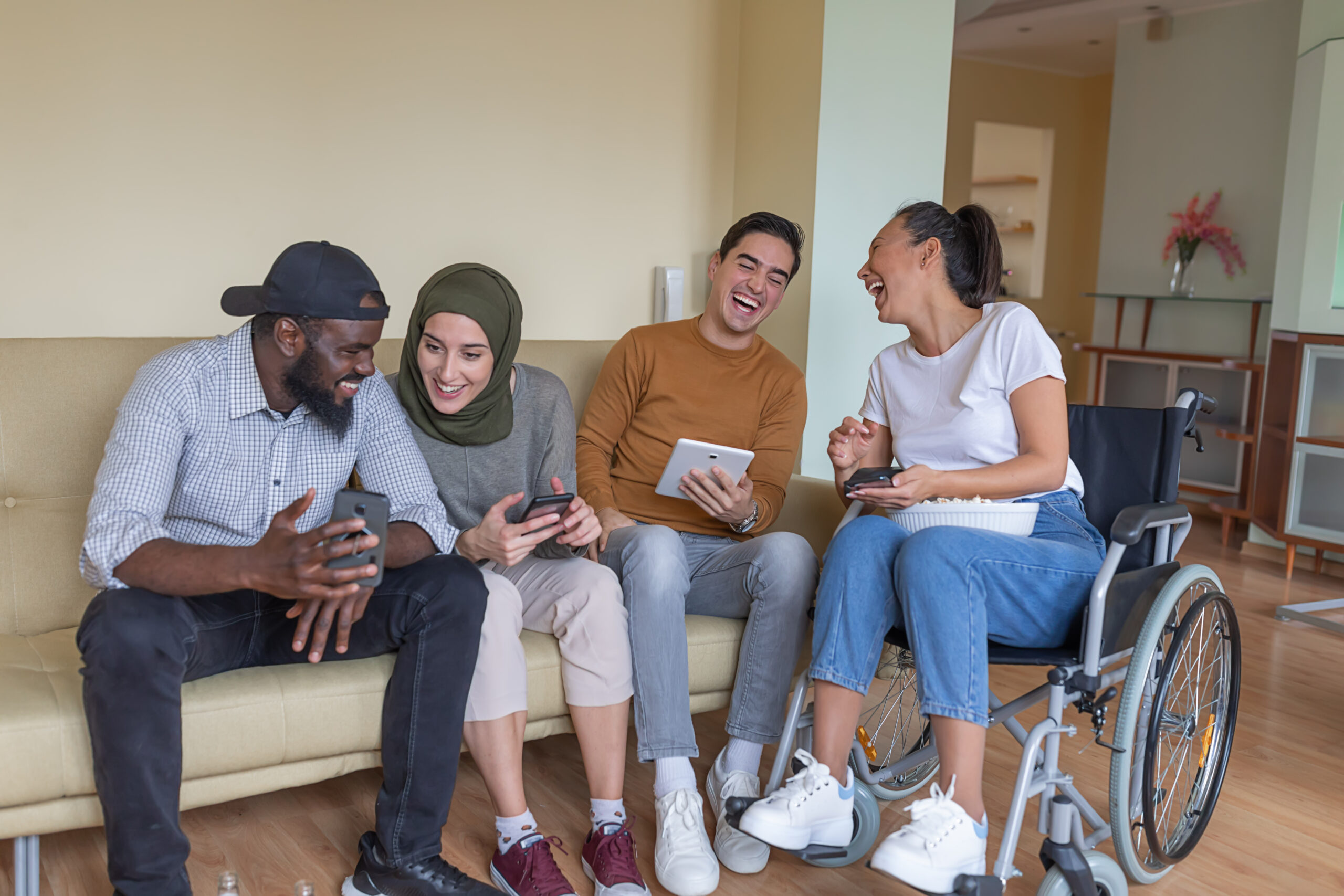 An Asian Woman in a Wheelchair and her Multi-Ethnic Young Friends are Spending a Nice Time Together in Their Comfortable Apartment, Hanging Out, Using Digital Devices, and Enjoys in Refreshing Drink.