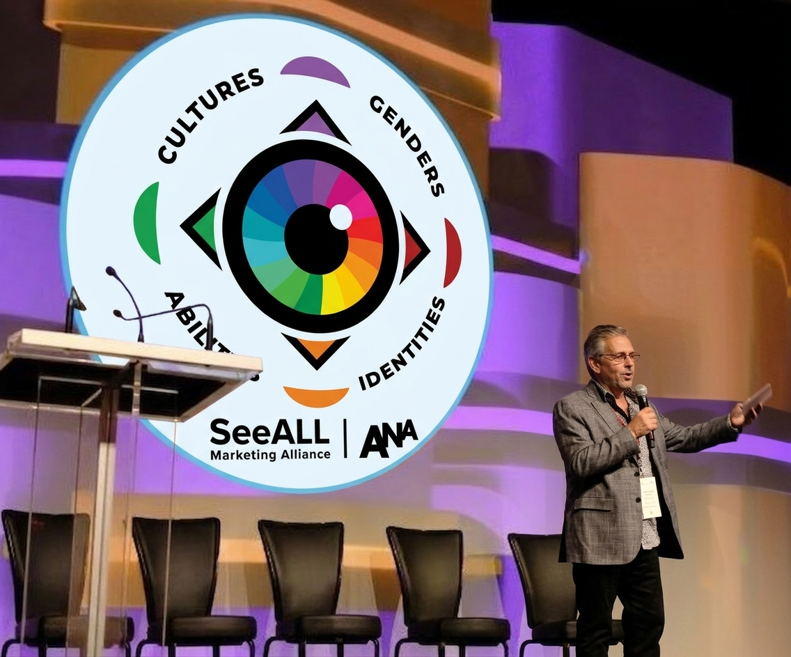 man on stage with 5 chairs behind him, a podium to the left and a logo on a screen in the background